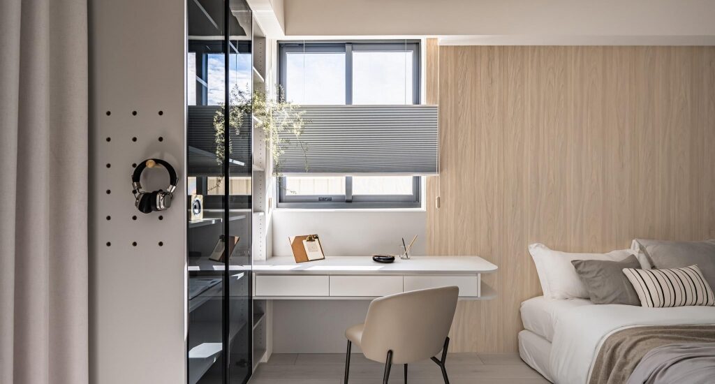 Modern bedroom with a white desk by a window, beige chair, and a wood-paneled wall; headphones hang on a pegboard to the left.